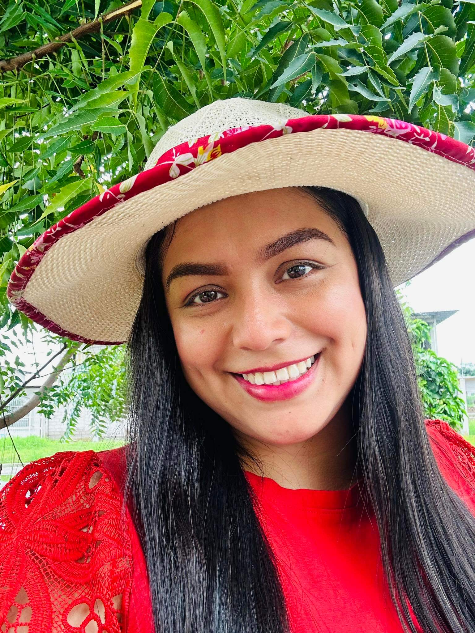Close-up of a smiling woman wearing a wide-brim straw hat with a red trim, green leaves in the background.